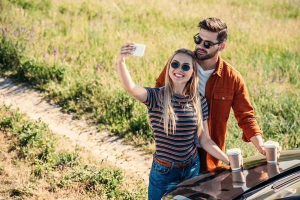 Elevated view of stylish couple in sunglasses with coffee cups taking selfie on smartphone near car on rural meadow — Stock Photo