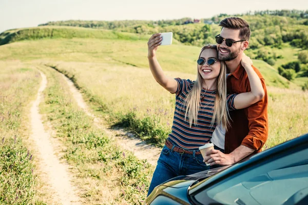 Sourire couple élégant dans des lunettes de soleil avec tasse à café prendre selfie sur smartphone près de la voiture sur prairie rurale — Photo de stock