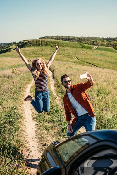 Sonriente joven saltando con los brazos anchos mientras su novio se toma selfie en el teléfono inteligente cerca del coche en el prado rural - foto de stock