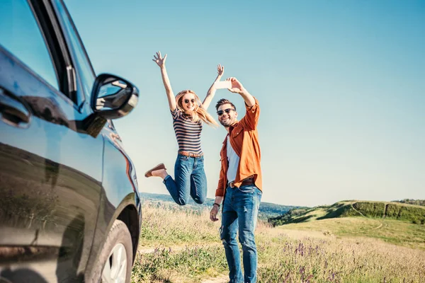 Feliz mujer joven saltando con los brazos anchos mientras que su novio tomando selfie en teléfono inteligente cerca de coche en el campo - foto de stock