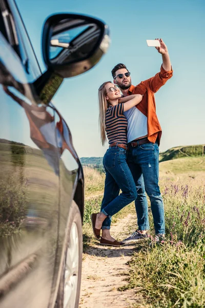 Homem elegante em óculos de sol tomando selfie no smartphone com namorada sorridente perto do carro no prado rural — Fotografia de Stock