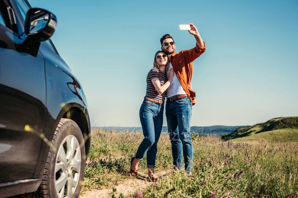 Homem elegante tomando selfie com namorada perto de carro no prado rural — Fotografia de Stock
