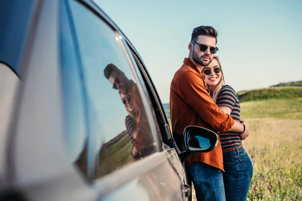 Smiling stylish couple in sunglasses standing near car on rural meadow — Stock Photo