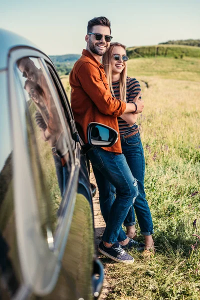 Стильний усміхнений пара в сонцезахисні окуляри, стоячи біля автомобіля на сільських Луці — стокове фото