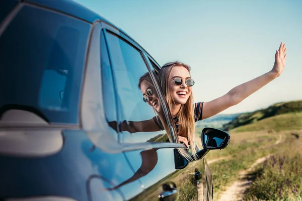 Stylish woman in sunglasses leaning out from car window and waving hand on rural meadow — Stock Photo