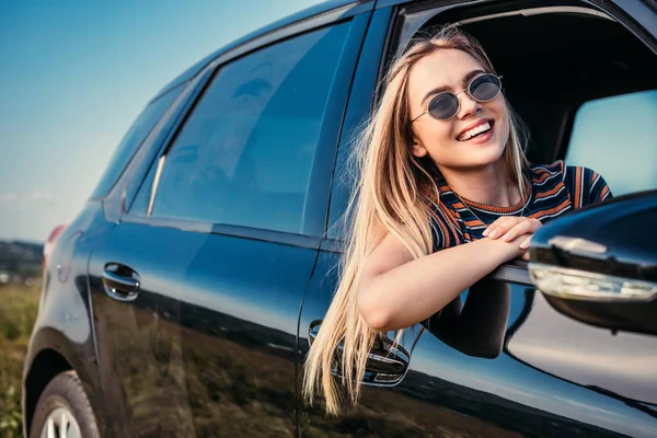 Щаслива стильна жінка в сонцезахисних окулярах, що виходить з вікна автомобіля — стокове фото