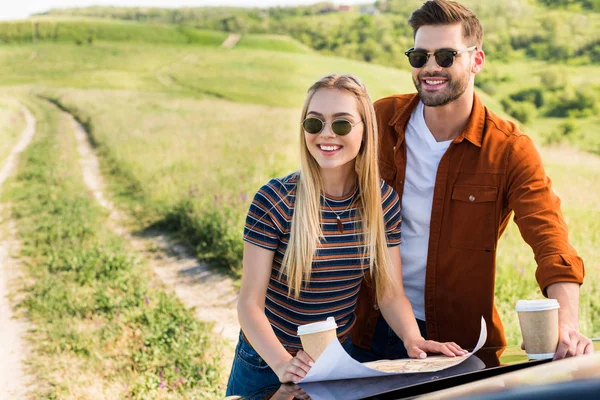 Sonriendo elegante pareja de viajeros con tazas de papel de café en busca de destino en el mapa cerca del coche en el campo rural - foto de stock