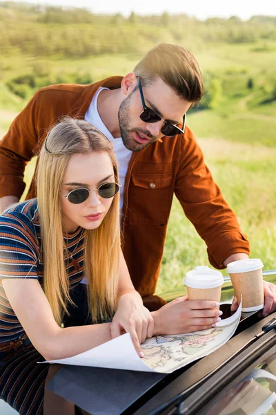 Stylish couple of tourists with paper cups of coffee looking for destination on map near car in rural field — Stock Photo