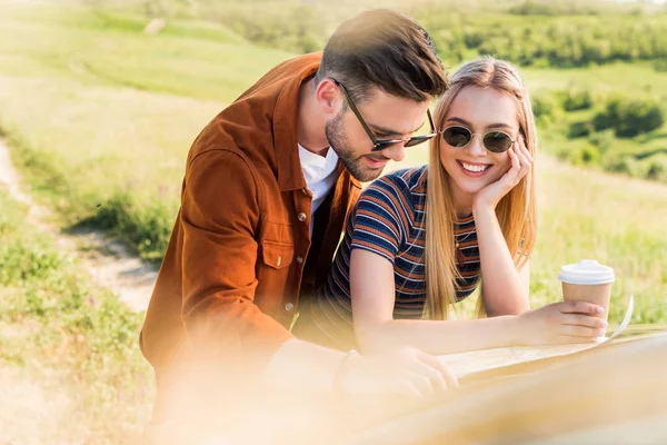 Feliz pareja de turistas con tazas de papel de café en busca de destino en el mapa cerca de coche en el campo rural - foto de stock