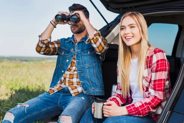 Turista masculino mirando a través de prismáticos mientras que su novia sonriente sentado cerca con taza de café en el maletero del coche en el campo - foto de stock