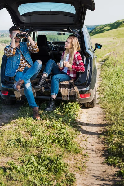 Stylish male tourist looking through binoculars while his smiling girlfriend sitting near with coffee cup on car trunk in field — Stock Photo