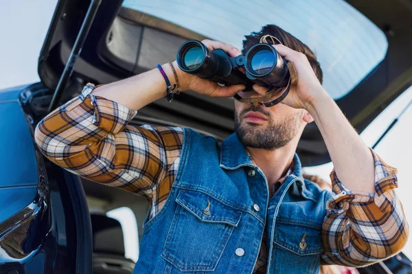 Vista basso angolo di turista maschio guardando attraverso binocoli mentre la sua ragazza seduta vicino sul bagagliaio auto — Foto stock