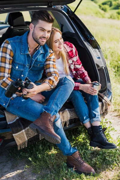 Männlicher Tourist mit Fernglas und seine lächelnde Freundin, die in der Nähe mit Kaffeetasse auf dem Kofferraum im Feld sitzt — Stockfoto