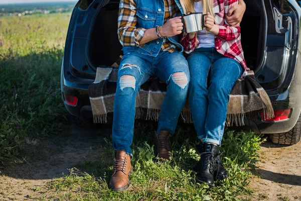 Tiro cortado de casal de turistas clinking por copos de café e sentado no porta-malas do carro no campo rural — Fotografia de Stock