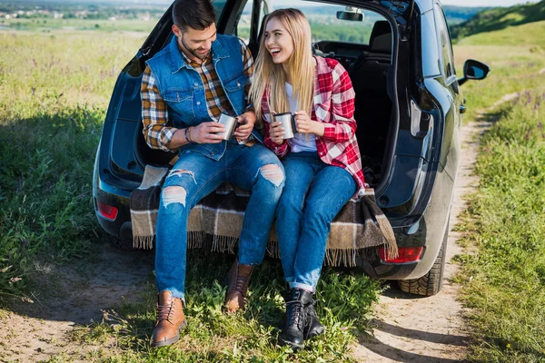 Feliz pareja con tazas de café sentado en el maletero del coche en el campo rural - foto de stock