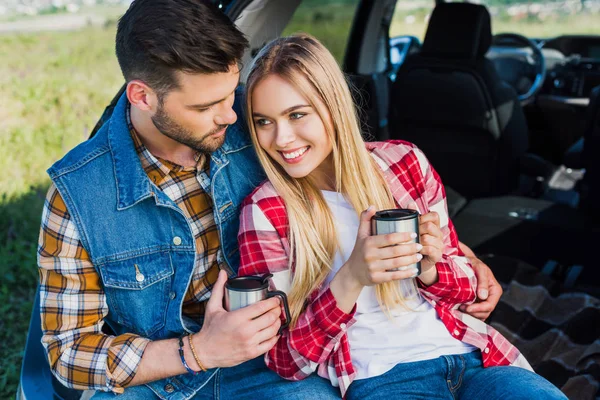 Jovem casal com xícaras de café sentado no carro tronco no campo rural — Fotografia de Stock