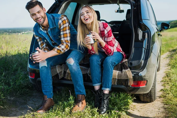 Smiling couple of stylish tourists with coffee cups sitting on car trunk in rural field — Stock Photo