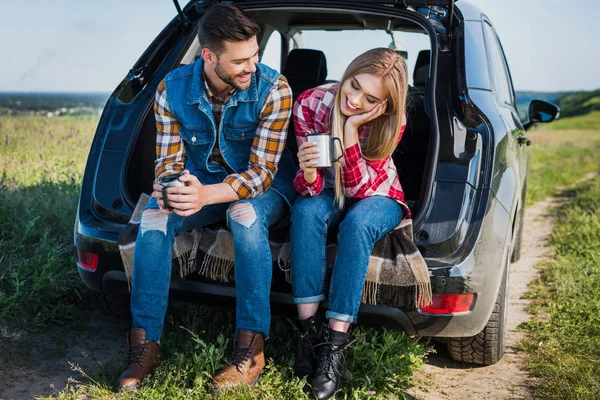 Pareja con tazas de café hablando entre sí y sentado en el maletero del coche en el campo rural - foto de stock