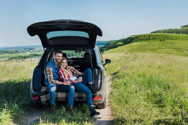 Happy couple of stylish tourists with coffee cups sitting on car trunk in rural field — Stock Photo