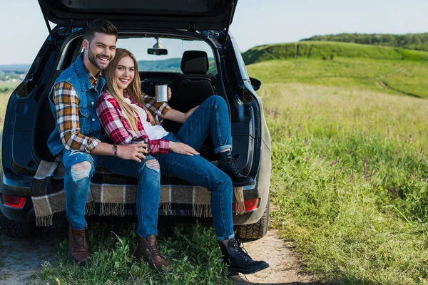 Sonriente pareja con tazas de café sentado en el maletero del coche en el campo rural - foto de stock