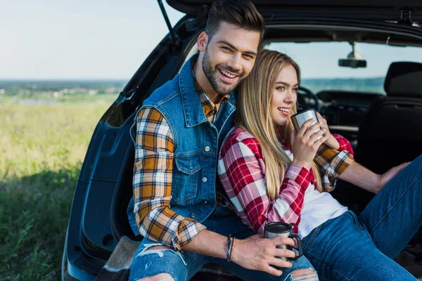 Smiling couple with coffee cups sitting on car trunk in rural field — Stock Photo