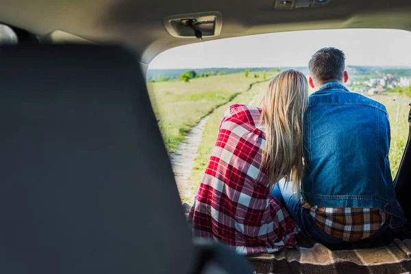 Back view of couple of stylish travelers sitting on car trunk in rural field — Stock Photo