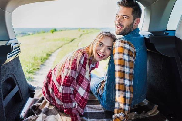Joven pareja sonriente sentado en el maletero del coche y mirando a la cámara en el campo rural - foto de stock