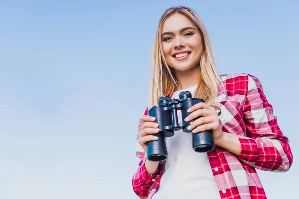 Улыбающаяся женщина-путешественница держит бинокль на фоне голубого неба — стоковое фото
