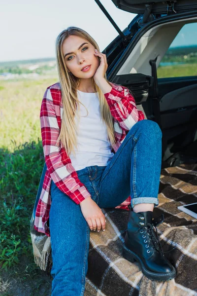 Smiling young woman sitting on car trunk in rural field — Stock Photo