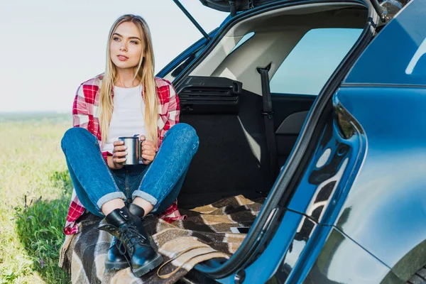 Молодая женщина сидит на багажнике автомобиля с чашкой кофе в сельской местности — стоковое фото