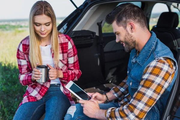 Uomo sorridente utilizzando tablet digitale con schermo bianco mentre la sua ragazza seduta vicino con tazza di caffè sul bagagliaio dell'auto — Foto stock