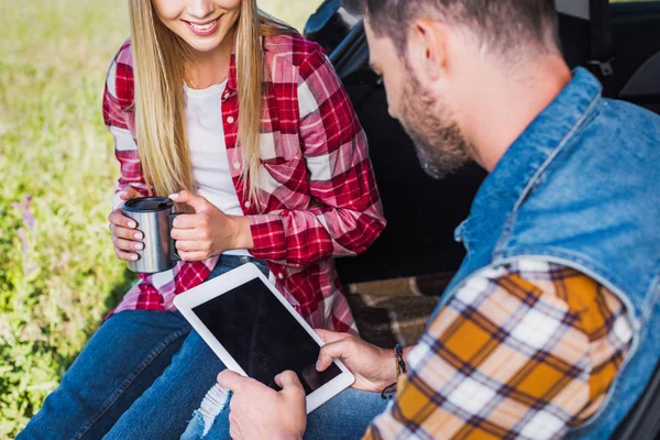 Abgeschnittenes Bild des Mannes mit digitalem Tablet mit leerem Bildschirm, während seine Freundin in der Nähe mit Kaffeetasse auf dem Kofferraum sitzt — Stockfoto