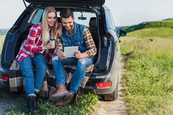 Щасливий чоловік використовує цифровий планшет, а його дівчина сидить поруч з чашкою кави на багажнику автомобіля — стокове фото