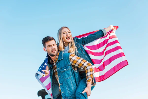 Stylish man doing piggyback ride to girlfriend holding american flag against blue sky, independence day concept — Stock Photo