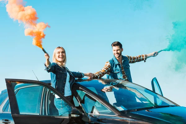 Smiling couple standing on car and holding green and orange smoke bombs — Stock Photo