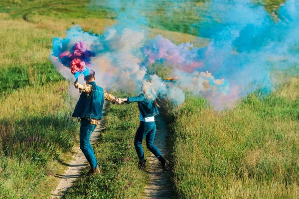 Back view of couple holding colorful smoke bombs in rural field — Stock Photo