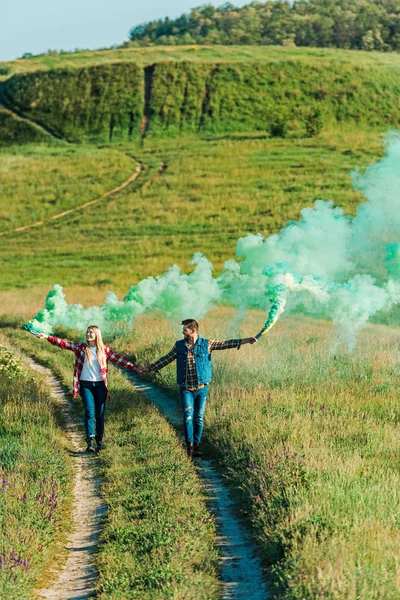 Vue surélevée d'un jeune couple tenant des bombes fumigènes vertes sur un pré rural — Stock Photo