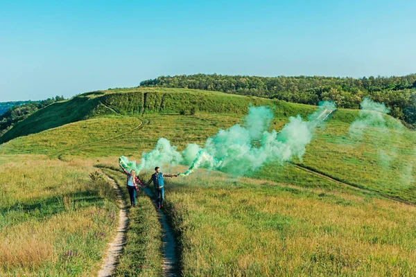 Vue lointaine d'un jeune couple tenant des bombes fumigènes vertes sur un pré rural — Photo de stock