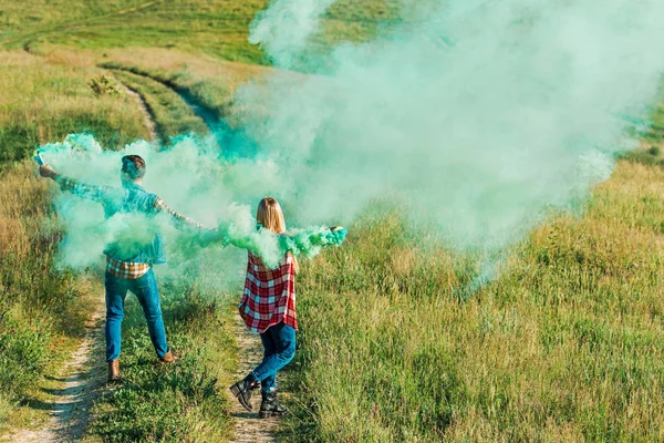 Back view of young couple holding green smoke bombs on rural meadow — Stock Photo