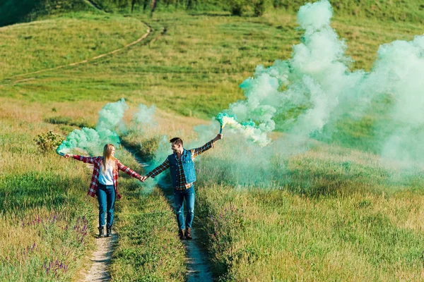 Jovem casal segurando bombas de fumaça verde no prado rural — Fotografia de Stock