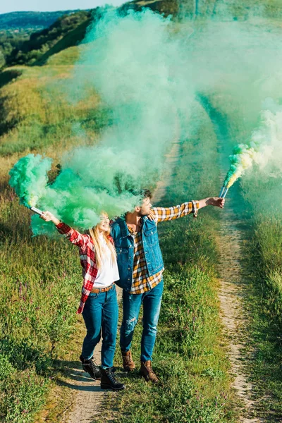 Vista elevada de pareja joven sosteniendo bombas de humo verdes en el campo - foto de stock