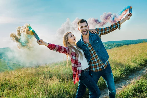 Joven pareja sosteniendo coloridas bombas de humo en prado rural - foto de stock