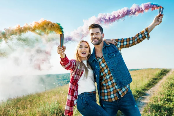 Smiling couple holding colorful smoke bombs on rural meadow — Stock Photo