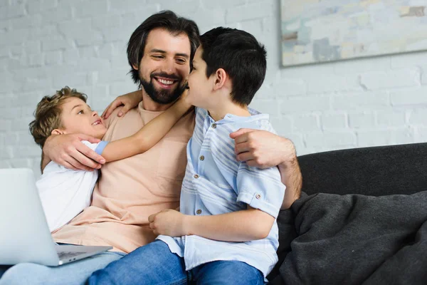 Retrato de homem com laptop e crianças perto abraçando uns aos outros no sofá em casa — Fotografia de Stock