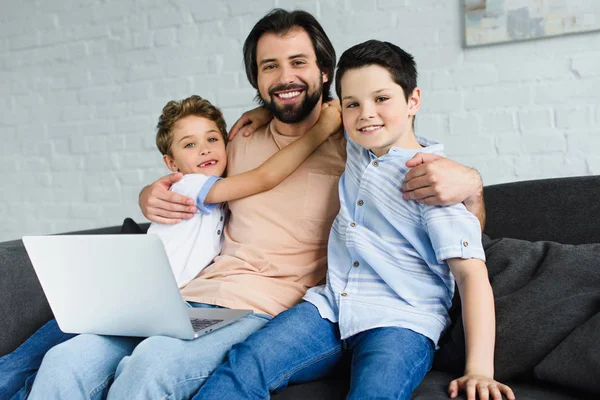 Retrato del hombre con el ordenador portátil y los niños cerca abrazándose en el sofá en casa - foto de stock