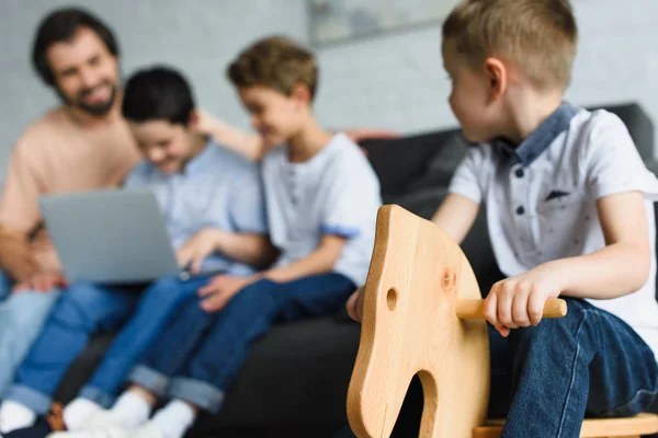 Selektiver Fokus des kleinen Jungen auf Holzpferd-Spielzeug und Familie mit Laptop zusammen auf dem heimischen Sofa — Stockfoto