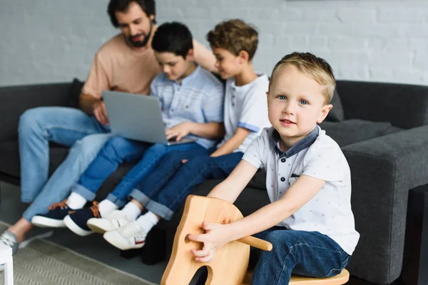 Selective focus of little boy on wooden horse toy and family using laptop together on sofa at home — Stock Photo
