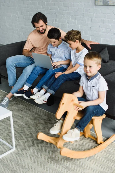 Homme et enfants à proximité en utilisant un ordinateur portable ensemble sur le canapé à la maison — Photo de stock