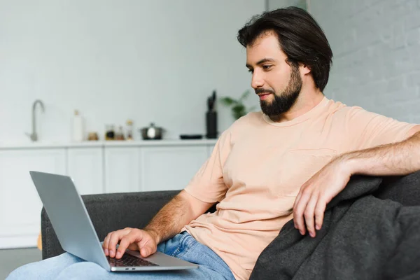 Side view of smiling man using laptop while resting on sofa at home — Stock Photo