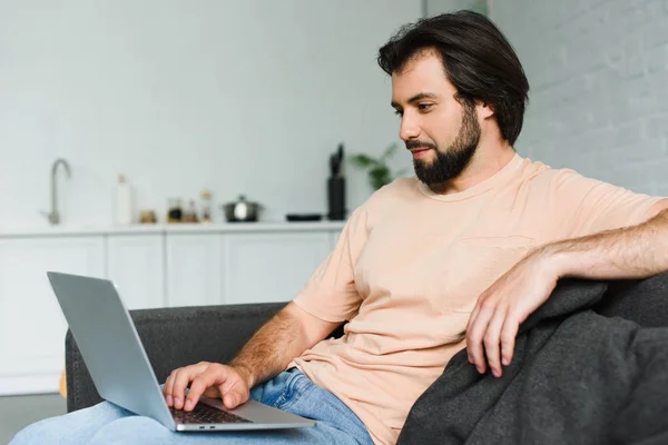 Vista lateral do homem sorridente usando laptop enquanto descansa no sofá em casa — Fotografia de Stock